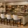 Modern custom wet bar with wood cabinetry, stone countertop, wine fridge, open shelving, glassware, and upholstered counter stools.