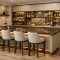 Modern custom wet bar with wood cabinetry, stone countertop, wine fridge, open shelving, glassware, and upholstered counter stools.
